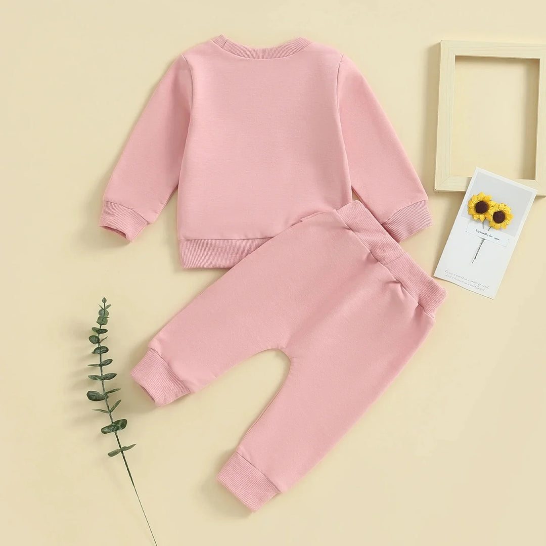 Soft pink long sleeve baby sweatshirt and matching pants on beige background with decorative flowers