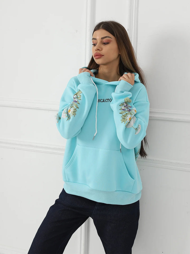 Woman wearing light blue hoodie with floral sleeve embroidery and kangaroo pocket posing against white wall