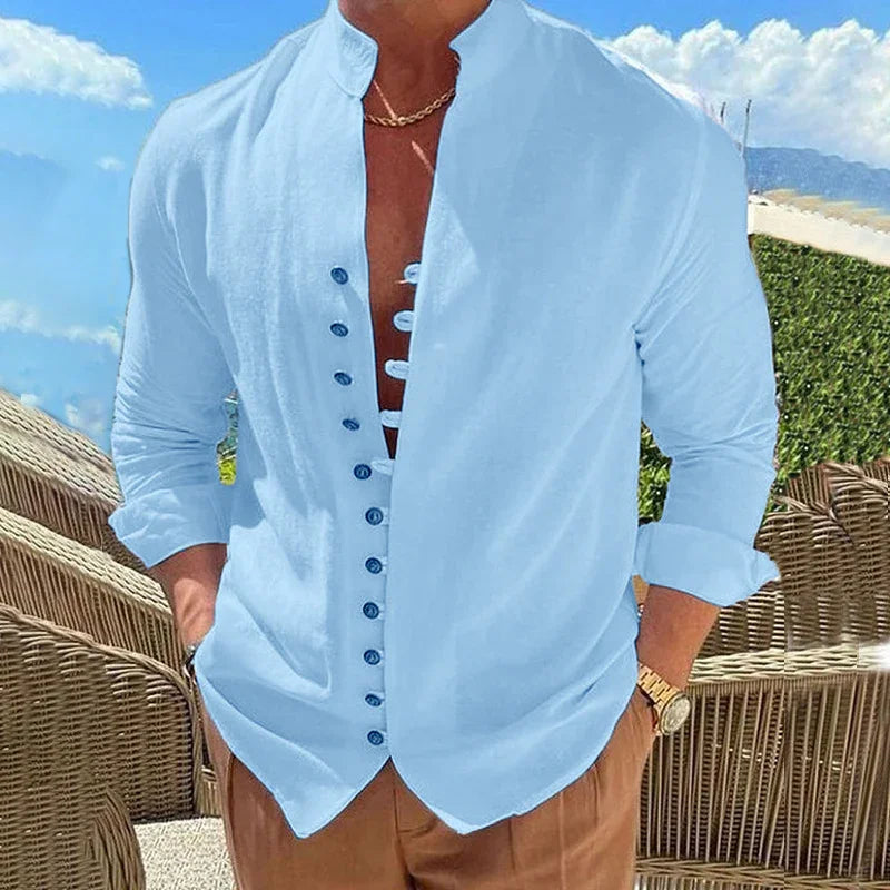 man wearing light blue buttoned casual shirt with rolled sleeves and brown pants outdoors with wicker chairs