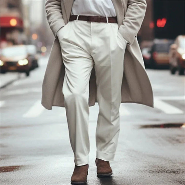 Man wearing beige trench coat, white pants, brown belt, and brown boots walking on city street