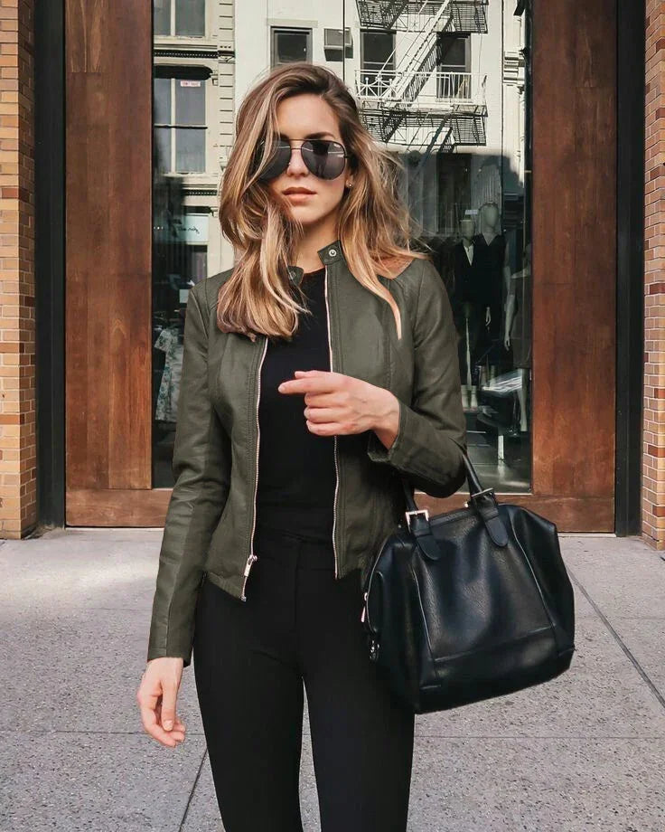 Stylish woman wearing black sunglasses, olive green leather jacket, and black outfit holding black handbag outside urban building