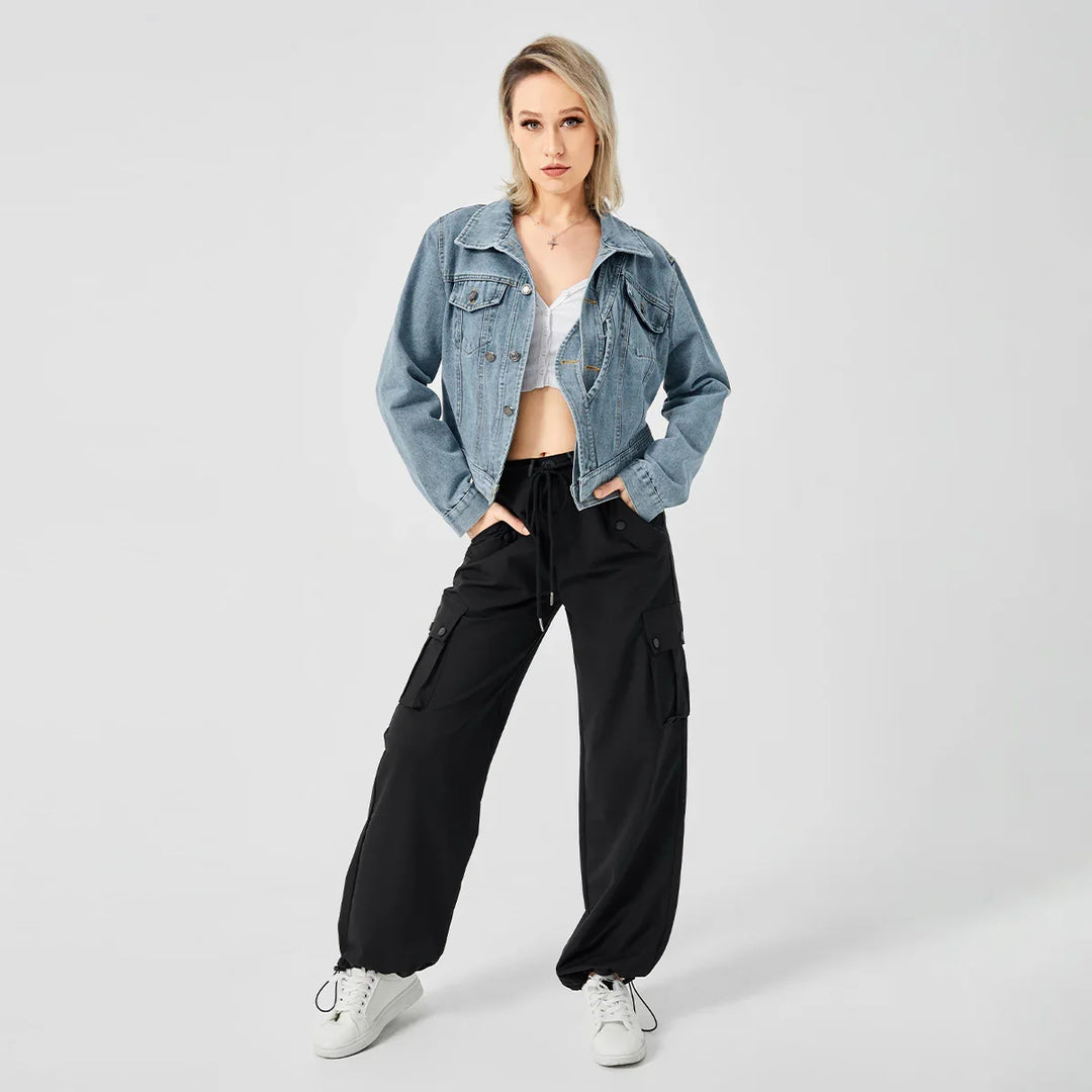 Blonde woman wearing light denim jacket, white crop top, black cargo pants, and white sneakers on gray background