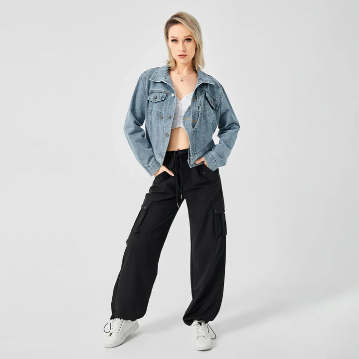 Blonde woman wearing light denim jacket, white crop top, black cargo pants, and white sneakers on gray background