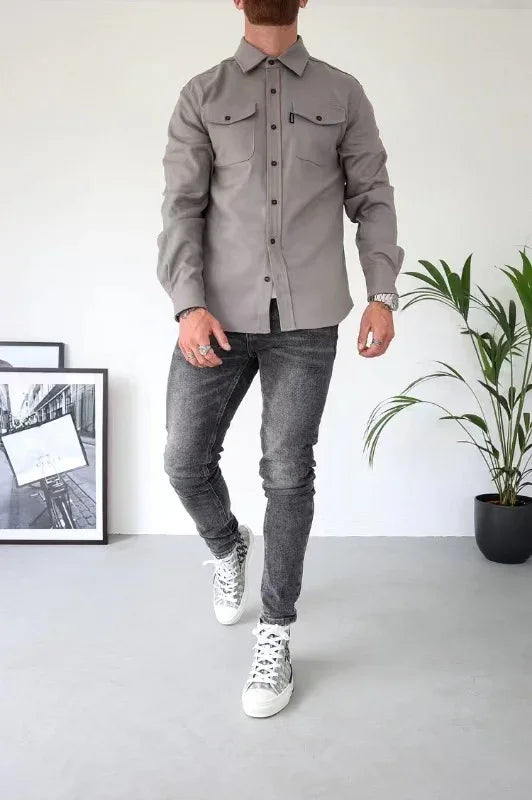 Man wearing gray button-up shirt, black jeans, and white sneakers standing indoors with a potted plant and framed photo