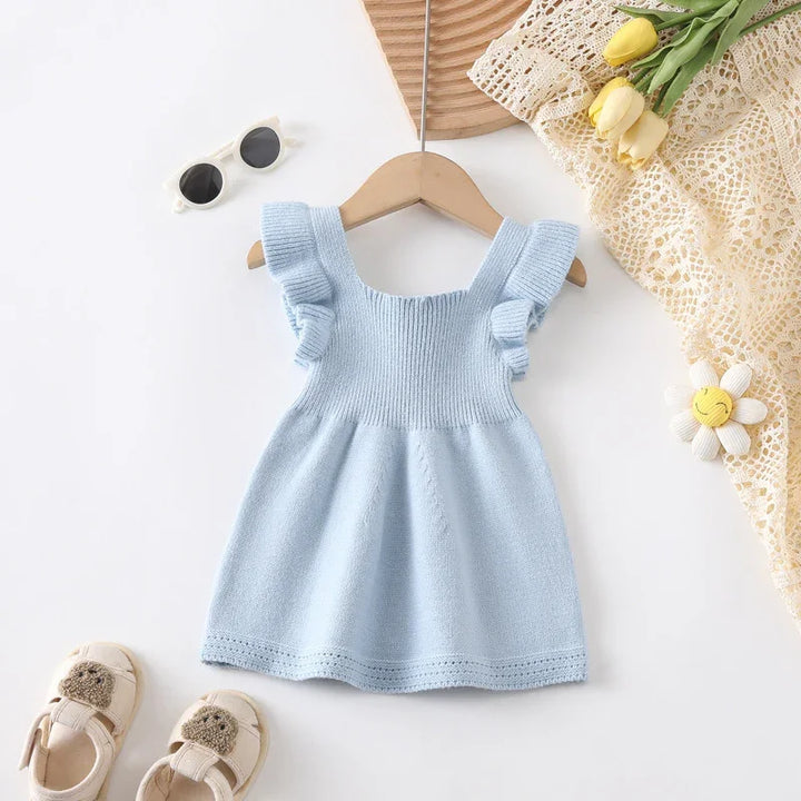 Light blue knitted baby dress with ruffled straps on wooden hanger with sunglasses, shoes, and yellow flowers