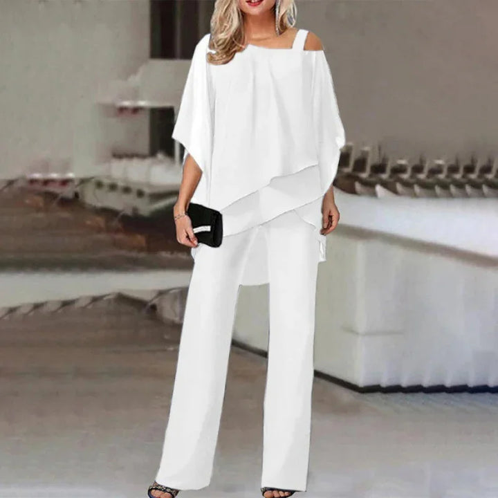 Woman wearing white asymmetrical top with shoulder straps and matching wide-leg pants holding black clutch