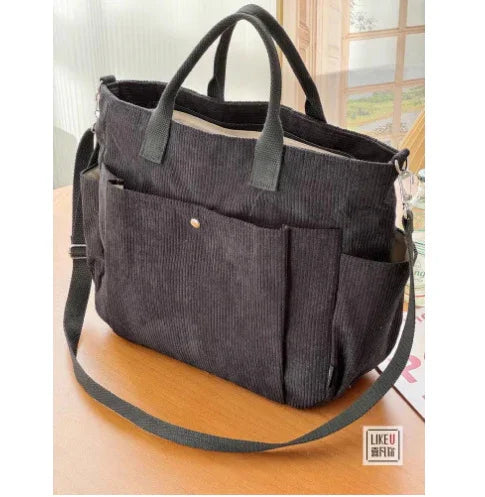 Black corduroy tote bag with front pocket, short handles, and adjustable shoulder strap on wooden table