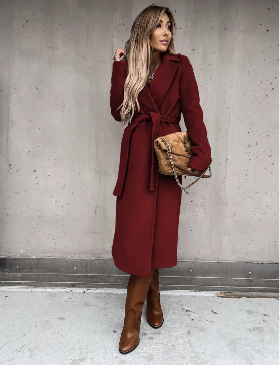 Woman in long maroon belted coat and brown boots holding tan quilted handbag against concrete wall
