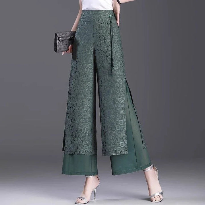 Woman wearing green floral lace wide-leg pants with side slits and sheer underlayer, styled with silver heels and clutch