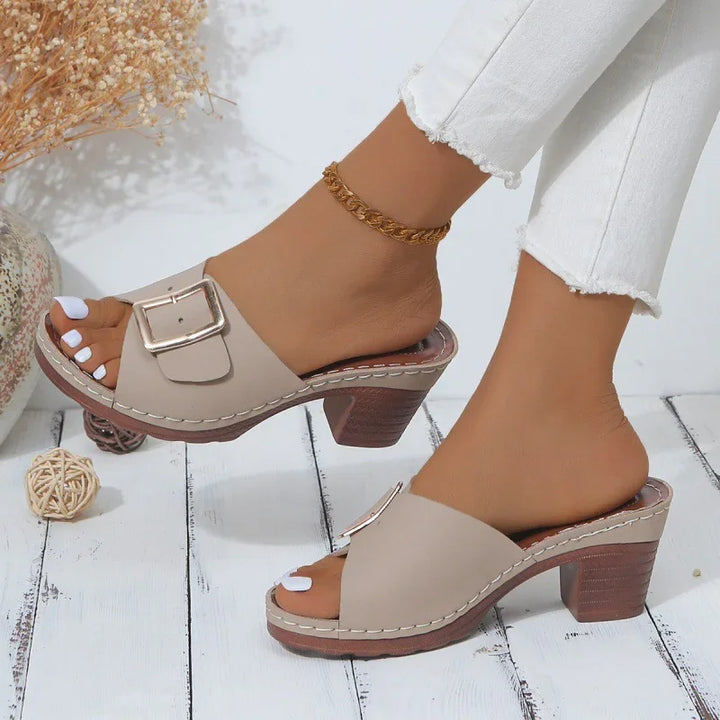 Beige heeled sandals with buckle detail on feet wearing white jeans and gold anklet on white wooden floor