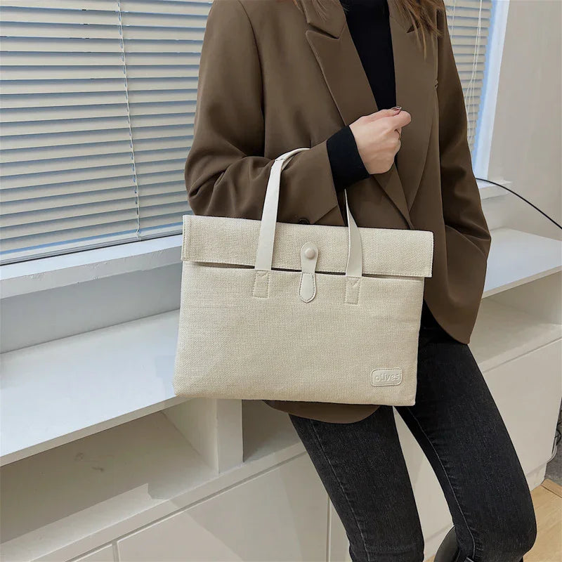 Woman holding beige canvas tote bag with button strap, brown blazer, black top, and dark jeans