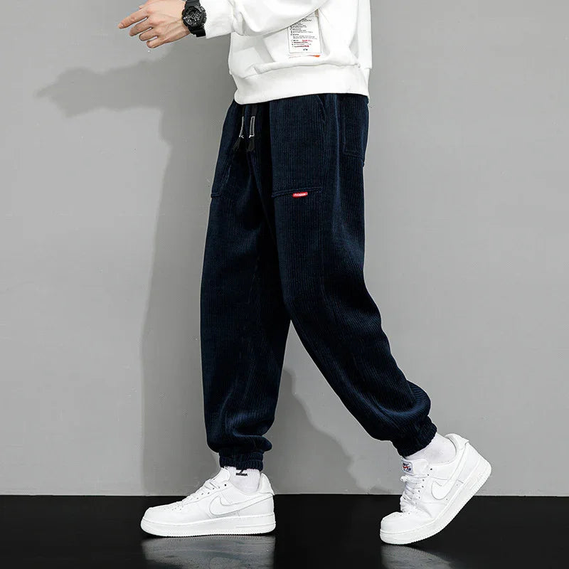 Man wearing black corduroy jogger pants with elastic cuffs and white sneakers walking against gray background