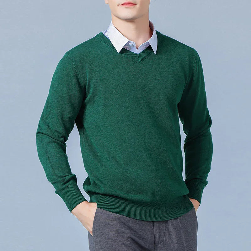 Man wearing dark green V-neck sweater over light blue collared shirt with gray pants against gray background