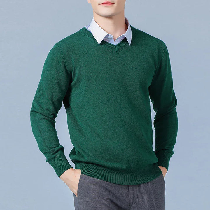 Man wearing dark green V-neck sweater over light blue collared shirt with gray pants against gray background