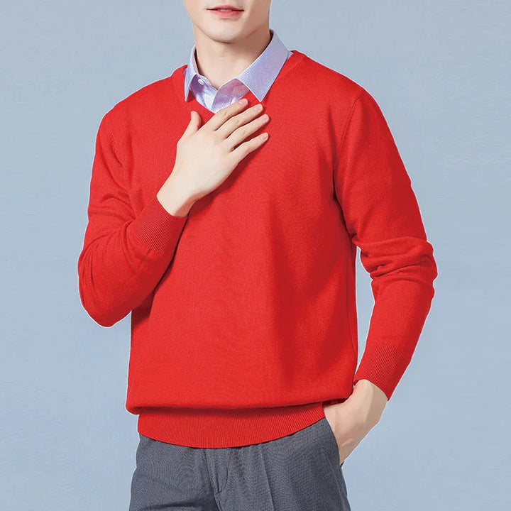 Man wearing red V-neck sweater over blue dress shirt with hand on chest and gray pants