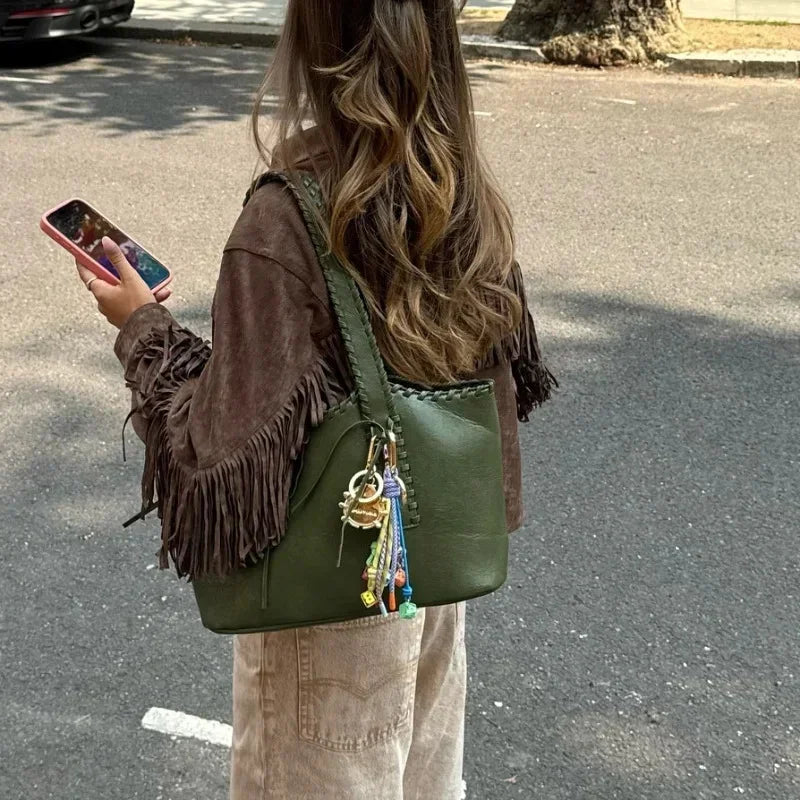 Woman with long wavy hair wearing brown fringe jacket and beige pants, holding green leather handbag with colorful charms outdoors