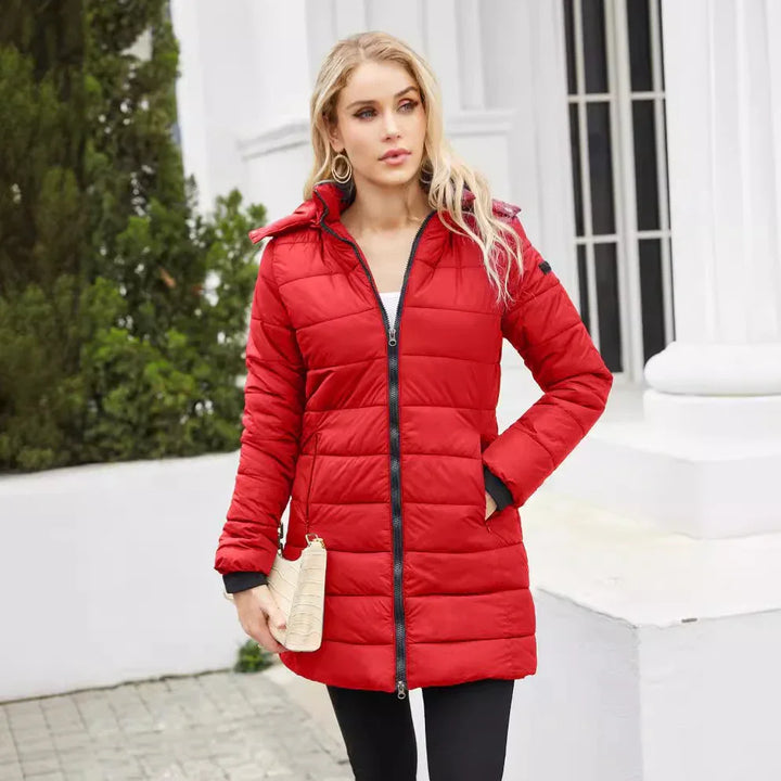Blonde woman wearing a red quilted winter coat standing outdoors near white building and greenery