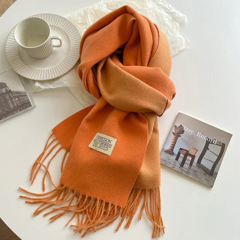 Orange gradient wool scarf with fringes on white table next to white cup and saucer and a photo book