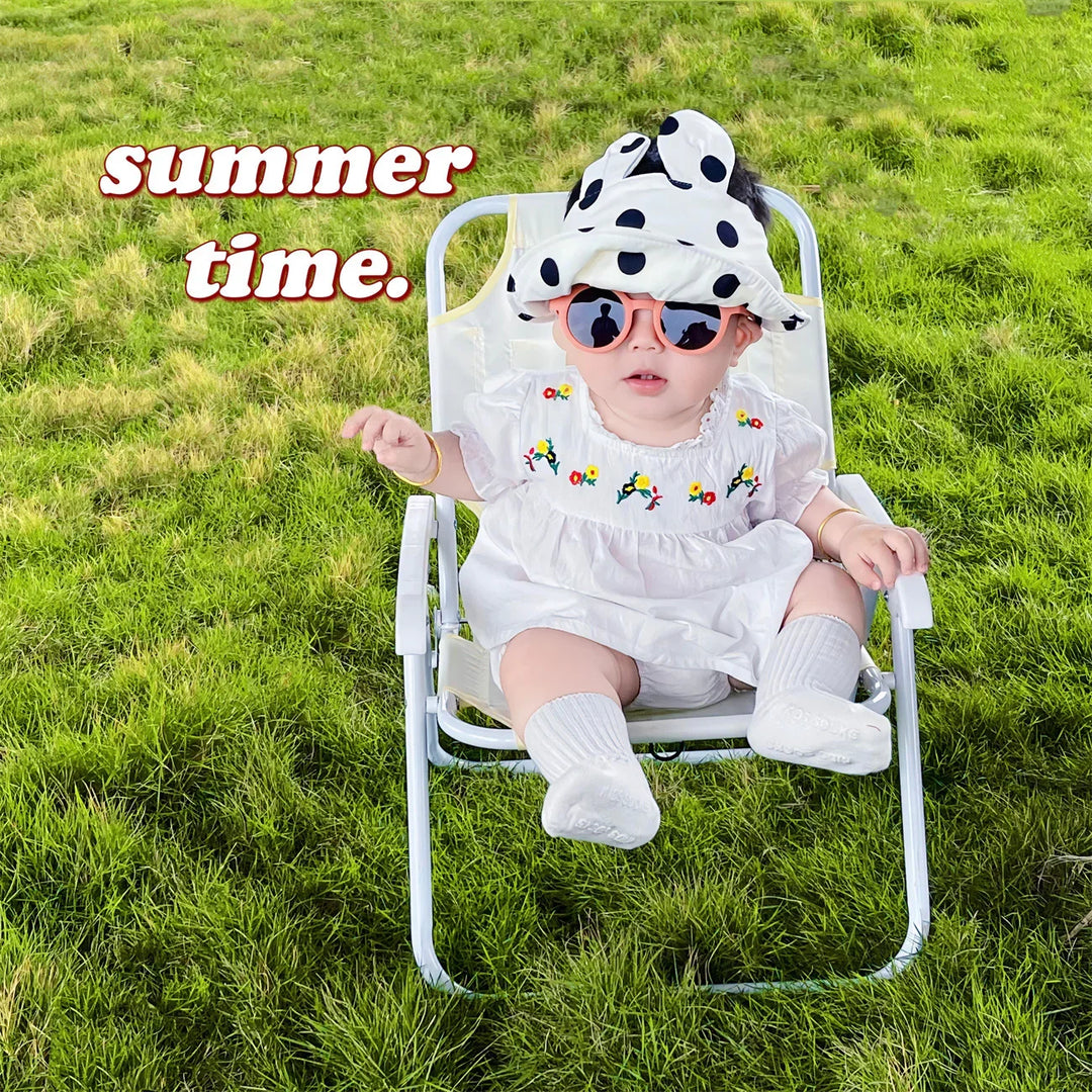 Baby in white dress and polka dot hat sitting on lawn chair on green grass with summer time text