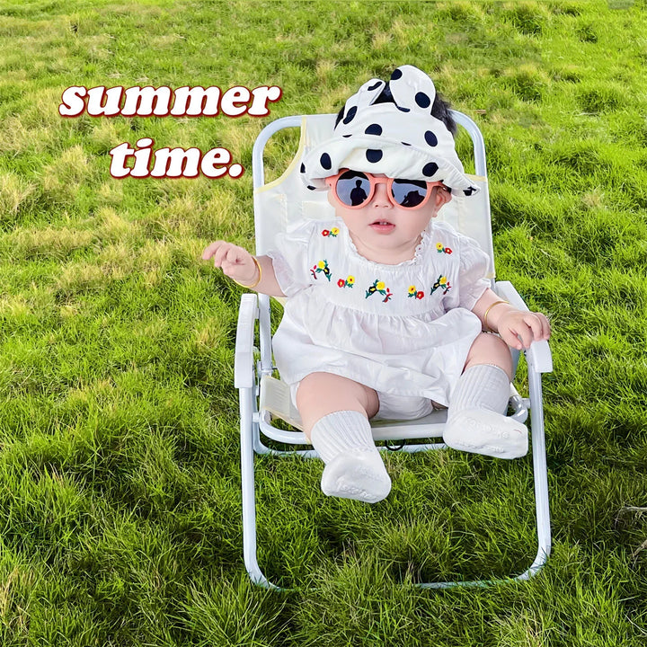 Baby in white dress and polka dot hat sitting on lawn chair on green grass with summer time text